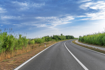 An empty, curved highway stretches through a lush countryside surrounded by tall grass and dense...