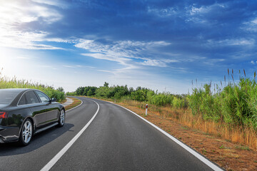A black sedan is parked on the side of a curved, tree-lined highway under a bright blue sky with...