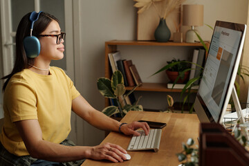 Side view shot of cheerful young Caucasian woman in headphones sitting at desktop computer, writing prompt to generate image online