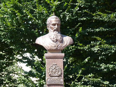  An Andrey Sheptytsky's bust with a cross necklace on a pedestal, against a backdrop of green foliage.