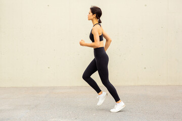 Fototapeta premium full length profile of sporty Asian woman running outdoors by urban wall