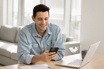 Fototapeta premium Handsome smiling man sitting at desk distracted from working on laptop using his modern smartphone, share messages, chatting, enjoy remote communication with friend, check email, reading pleasant news