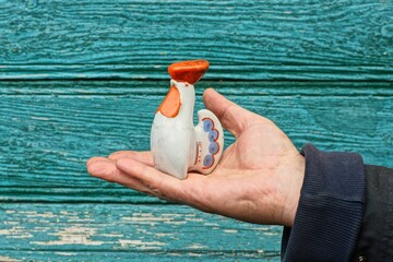 one small ceramic with krystalic salt kitchen beautiful closed white bottle in the form of a rooster with a red comb and tail with patterns in the hand of a man near a green wooden wall