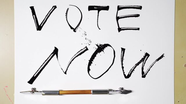 Hand revealing vote now message in bold black brush strokes on white paper.