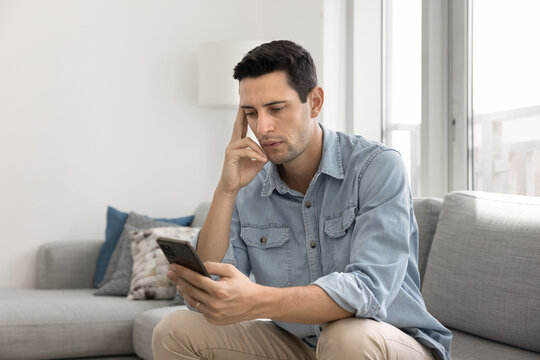 Young 30s Latin man read message with serious or concerned expression, get sms with news, review notification or e-mail from bank, using modern smartphone seated on couch in living room. Tech, apps
