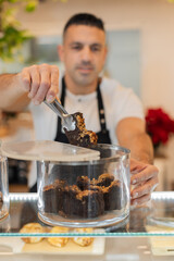 Close-up of chef using tongs to pick a walnut brownie from a transparent jar. Glass counter and...