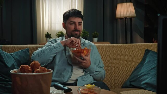 Bearded man spending time at home with fast food delivery, sits on couch watching tv and eating potato chips from a bowl. Slow motion