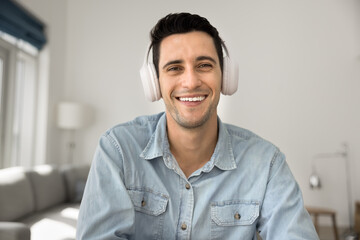 Smiling Hispanic man wear wireless headphones looking at camera record videovlog, take part in...