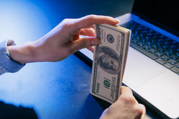 Hands holding a stack of US dollars near a laptop, symbolizing online earnings, digital finance, or internet business transactions