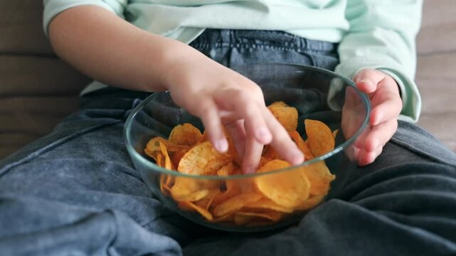 Child sitting at sofa and eating potato chips from a bowl.Caucasian kid have a junk food.Unhealthy eating.