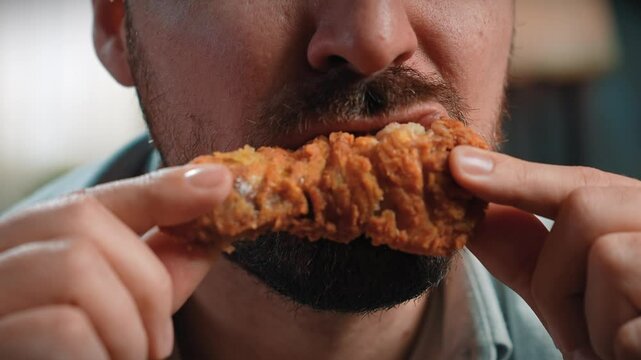 Close up shot of bearded man face man holding and biting chewing fried chicken leg, enjoying eating fast food. Slow motion
