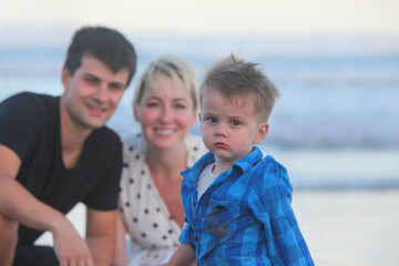 closeup portrait of sad serious toddler with his cheerful family in background on the beach at dusk 
