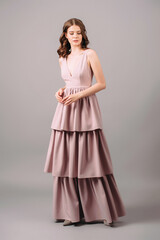 Fototapeta premium Romantic portrait of brunette woman in powder evening dress walking in studio. Bridesmaid's fashion. Dust pink plunge neckline sleeveless maxi gown with tiered skirt.Summer feminine look for hen party