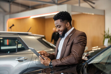 Young black car dealer standing at car showroom with tablet in hands and scrolling on it while...