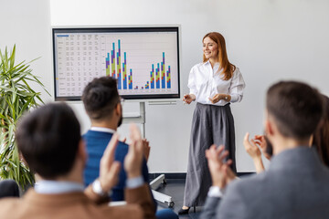 Data analytic expert standing by panel with charts and graphs and presenting it to her team in conference room.