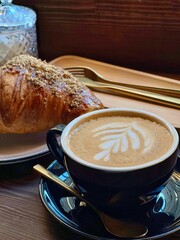 Cup of coffee, croissant and golden cutlery on wooden surface, traditional french breakfast photo in dark key