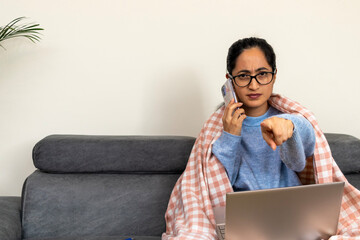 Young woman wearing soft blanket, working remotely on laptop while gesturing angrily during heated phone conversation, expressing workplace stress at home