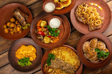 Traditional Eastern European cuisine served on rustic clay plates. Variety of homemade dishes including meat, potatoes, pancakes, dumplings, and grilled vegetables, on a wooden background