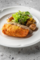 A beautifully plated dish featuring a golden-brown, crispy croquette served with roasted potatoes and a fresh salad, drizzled with a rich sauce on a textured stone surface.