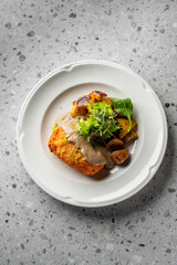 A beautifully plated dish featuring a golden-brown, crispy croquette served with roasted potatoes and a fresh salad, drizzled with a rich sauce on a textured stone surface.
