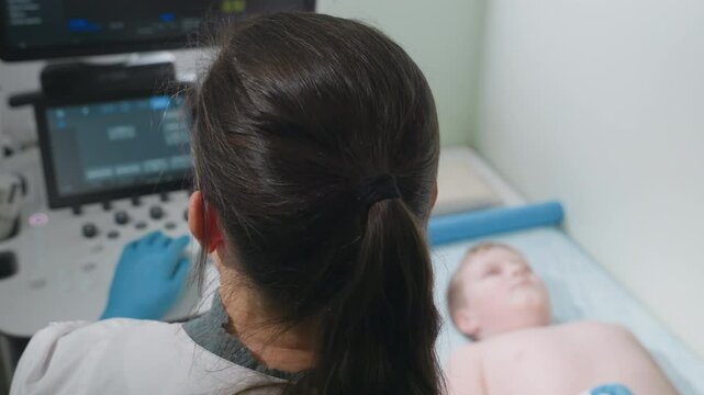 Aerial view of lab scientist with dark ponytail operating ultrasound machine while focused on screen displaying diagnostic data as barechested boy lies faintly in background on patient bed