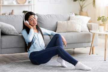 Obraz premium Relaxing Playlist. Young Asian Girl Listening Music In Wireless Headphones At Home, Sitting With Closed Eyes On Floor In Living Room, Free Space