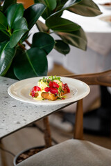 A beautifully plated dish featuring eggs Benedict topped with prosciutto, garnished with herbs, served on a stylish table with large green leaves in the background.
