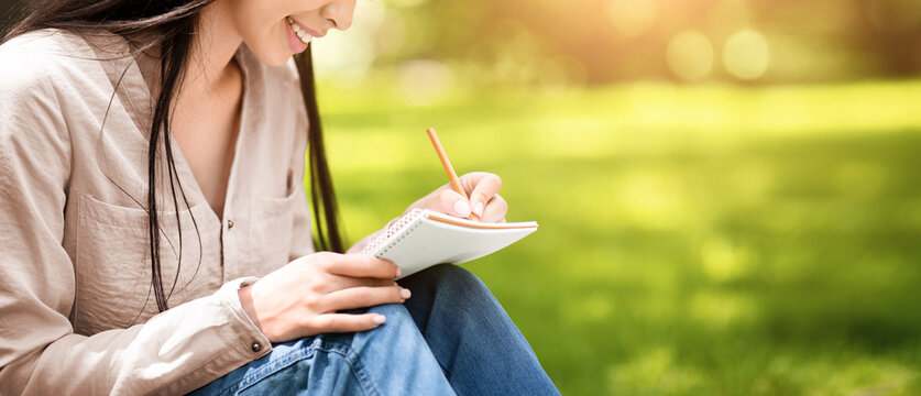 Young asian student girl writing notes to notebook while sitting on lawn in summer park, closeup shot with selective focus and copy space