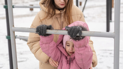 Young mother and a little girl in warm outerwear walk in the park in the children's playground in...