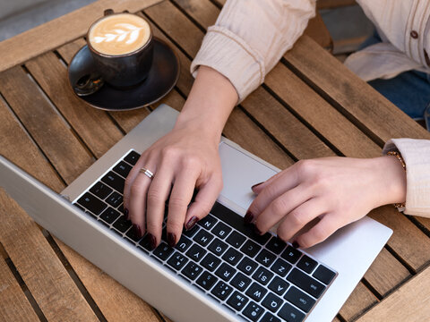 Close-up of female hands typing on laptop at café table. Casual outfit, cup of latte, remote work or digital nomad lifestyle.