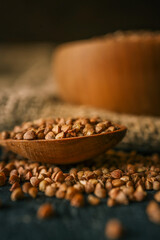 Spoonful of raw buckwheat groats on a dark surface with a wooden bowl and rustic burlap in the background. Healthy, natural food concept in warm earthy tones
