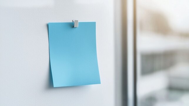 Single blue sticky note hanging on a white wall with a silver clip. the note is rectangular in shape and appears to be blank.