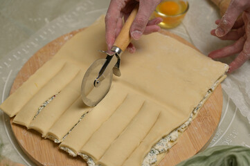 Close up square sheet of dough cut into strips with woman's hands on wooden board. Cutter, egg on kitchen table. Home cooking baking puffs
