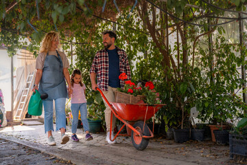 Gardeners walking out of a greenhouse, carrying vibrant flowers in a wheelbarrow and a watering...