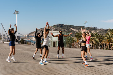 A group of six athletes jumping with their arms raised while looking at each other, outdoors,...