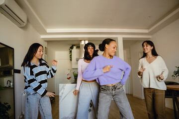 A group of three female friends stand and smile while looking at a female friend dancing in front...