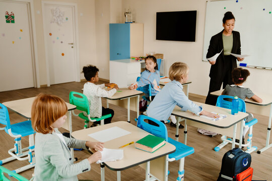 A teacher stands and talks while holding sheets of paper, a group of five schoolchildren sit at desks next to her, at school, in a classroom