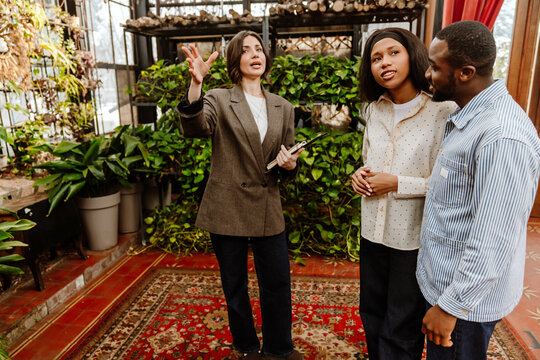 A man and a woman stand and listen to a female organizer who stands next to them and holds a clipboard while pointing in front of her, in a restaurant, wedding preparations