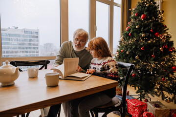 Grandfather and grandson sitting at the table and reading a book in front of them, in the living room, during Christmas