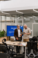 A male worker stands and talks with a male worker standing next to him and looking at the clipboard he is holding, in the hall, during retirement
