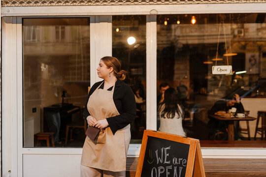 The female owner is standing and looking away, on the street at the entrance to the cafe