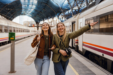 Fototapeta premium Two female friends walking and laughing while holding each other's hands and looking to the sides, at a train station, during a trip