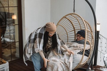 Grandma stands and covers grandfather with a blanket, who is sitting in a cocoon chair and smiling, outside, in winter