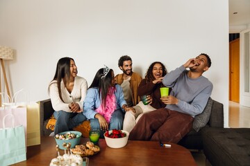 A group of five friends sitting on a sofa and laughing while looking at one of them, at a birthday party