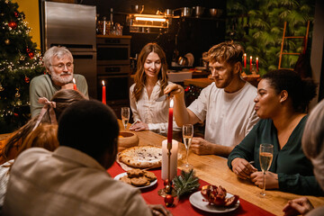Family and guests sitting at a table and looking at a man sitting between them and holding a knife during Christmas dinner