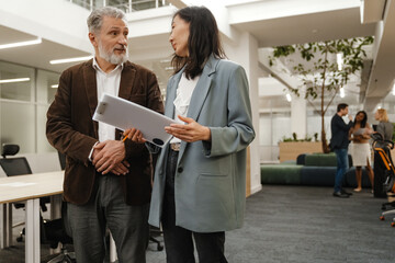 A male worker is walking and talking with a female worker who is walking next to him and holding a clipboard, in the hall, during retirement