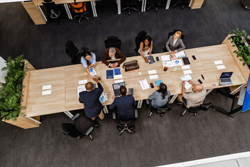 Top view of a group of eight workers sitting at a table, two of them shaking hands, in a hall, during retirement