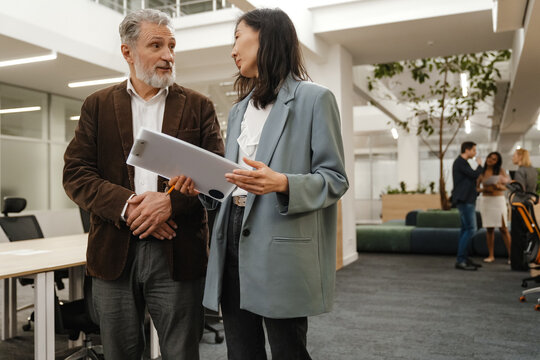 A male worker is walking and talking with a female worker who is walking next to him and holding a clipboard, in the hall, during retirement