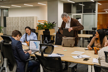 A White elder male office worker and his multinational group of colleagues collaborating in the office, with laptops and documents on the table, preparing for the next task in a modern workspace.
