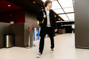 A White young man with glasses and beige headphones walking through a modern office lobby, carrying...